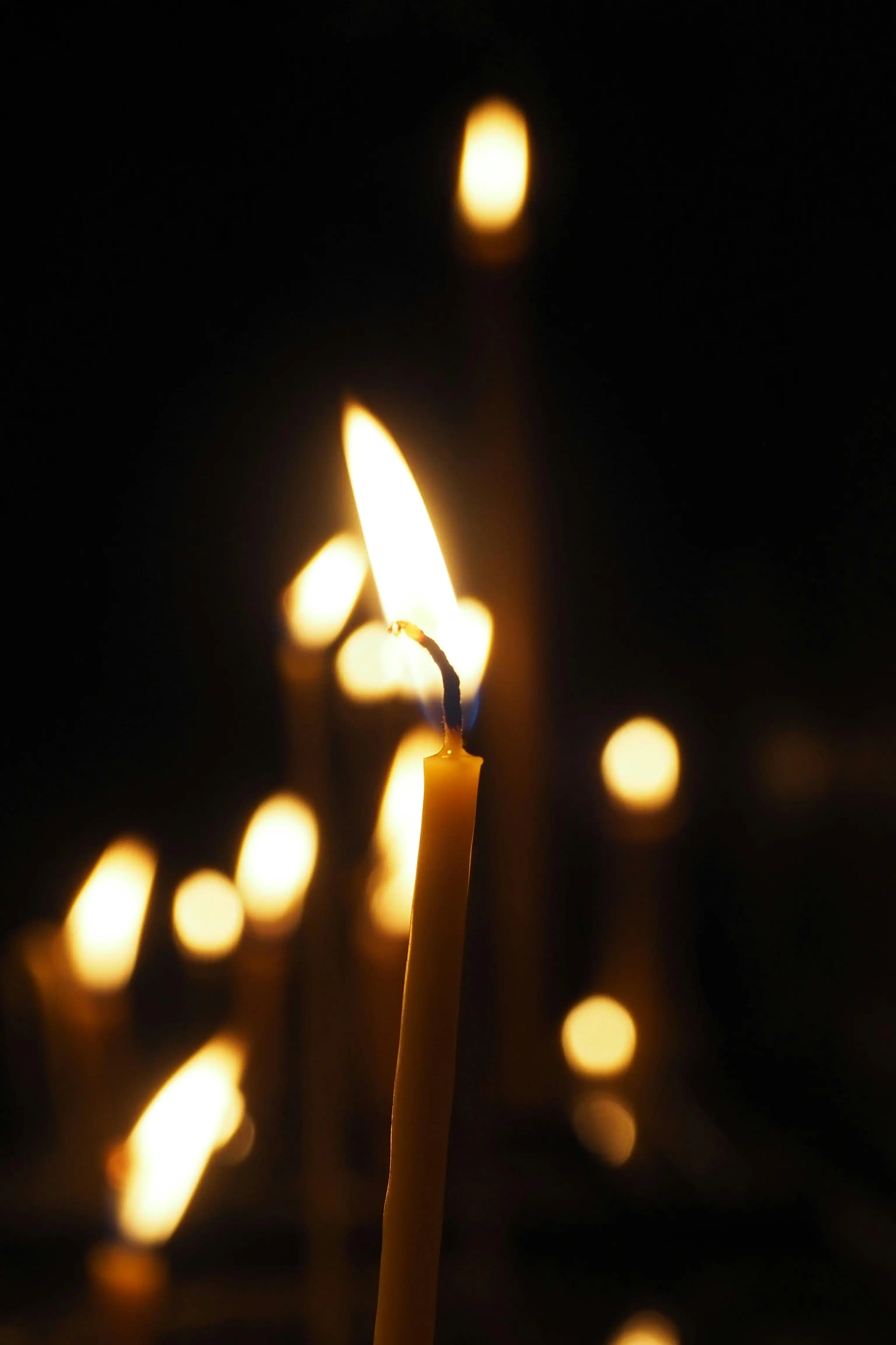 Tuvplānā degoša šaura vaska svece tumsā, fonā redzamas izplūdušas citu sveču liesmas enerģētiskās attīrīšanas rituālā.