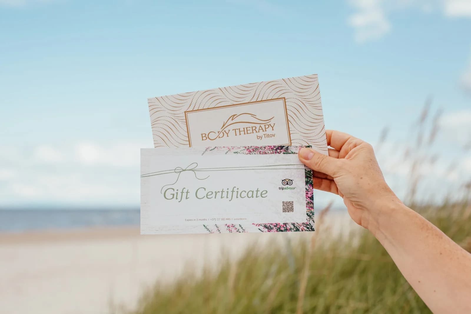 Коуч и массажный терапевт Алексейс Титов держит подарочные сертификаты Therapy by Titov на берегу моря в Риге, на фоне голубого неба и пляжа.