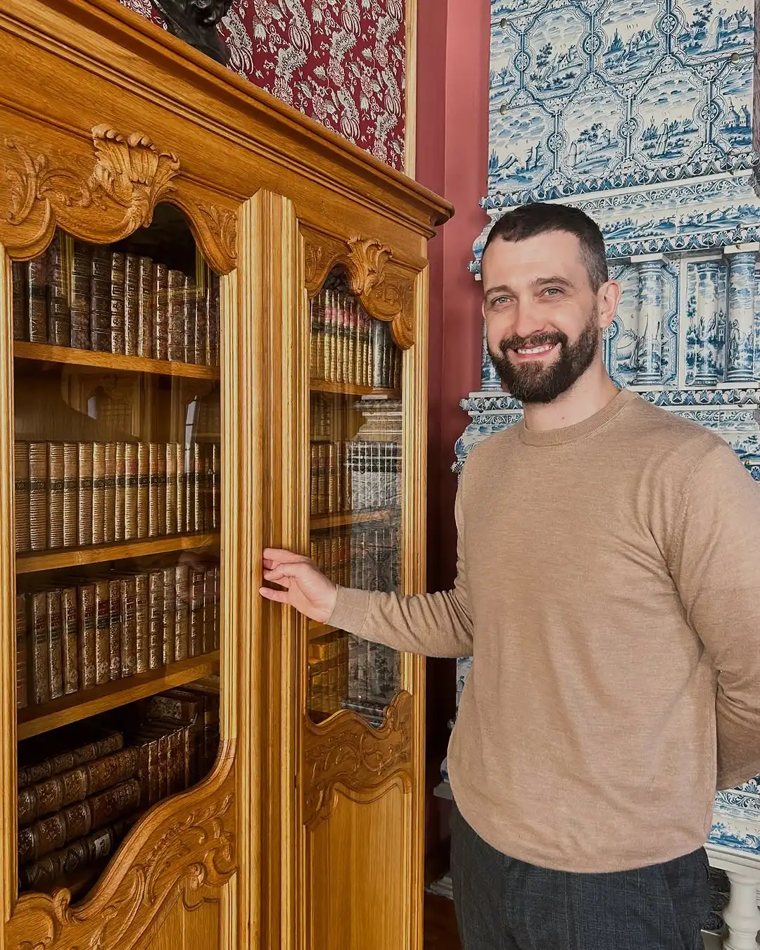 Aleksejs gaišā džemperī pie senlaicīga koka grāmatu skapja vēsturiskā interjerā.