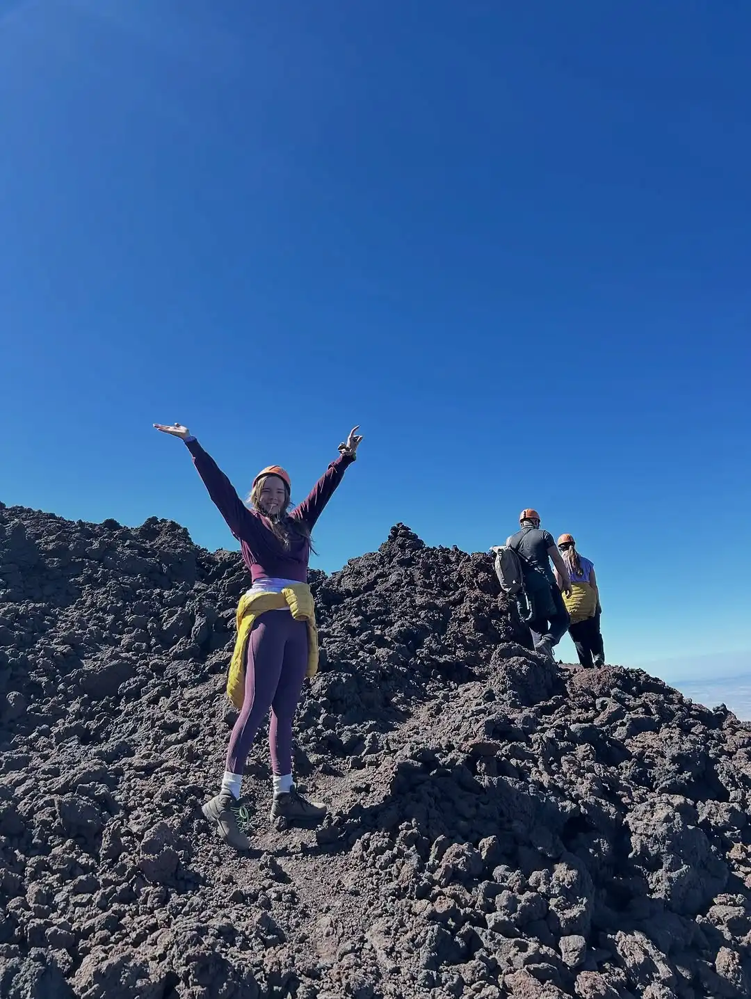Darja ar paceltām rokām stāv uz tumšiem vulkāna akmeņiem kalna virsotnē, blakus redzami citi kāpēji.