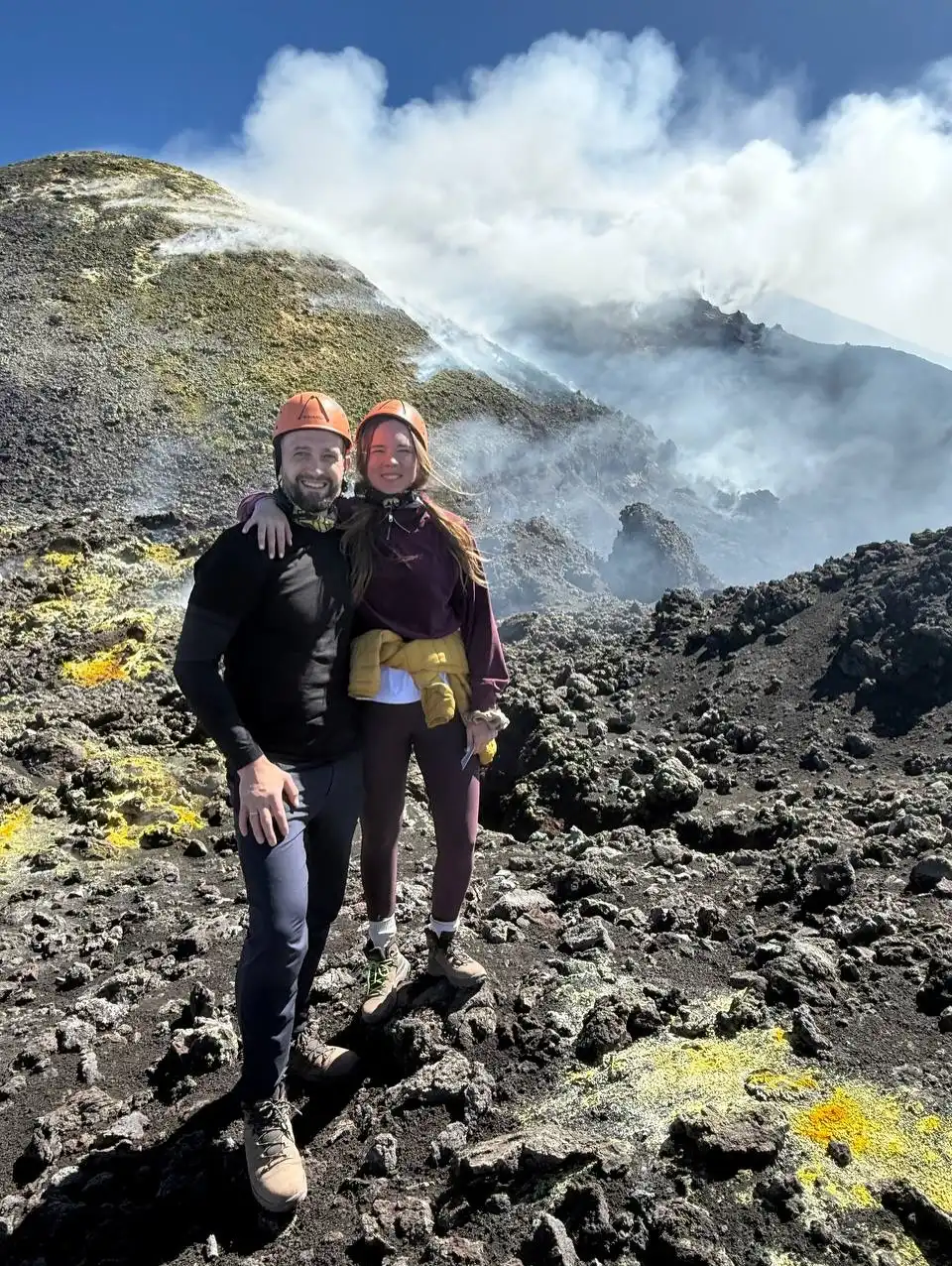 Aleksejs un Darja oranžās ķiverēs stāv uz melnas lavas pie dūmakaina vulkāna nogāzes.